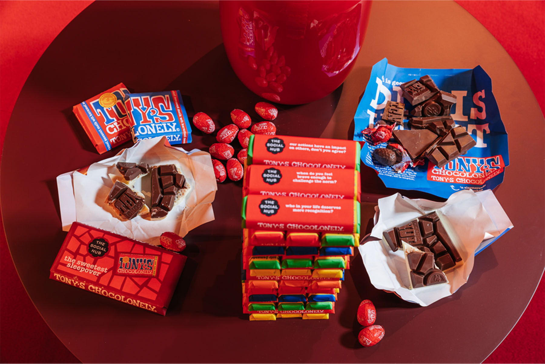 A display of Tony’s Chocolonely chocolate bars and mini eggs arranged on a table, some of the bars partially unwrapped. The wrappers feature co-branded packaging with The Social Hub logo visible