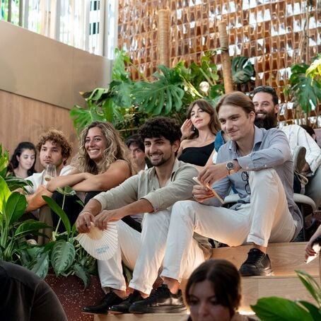 Audience seated on steps attending indoor event surrounded by lush plants