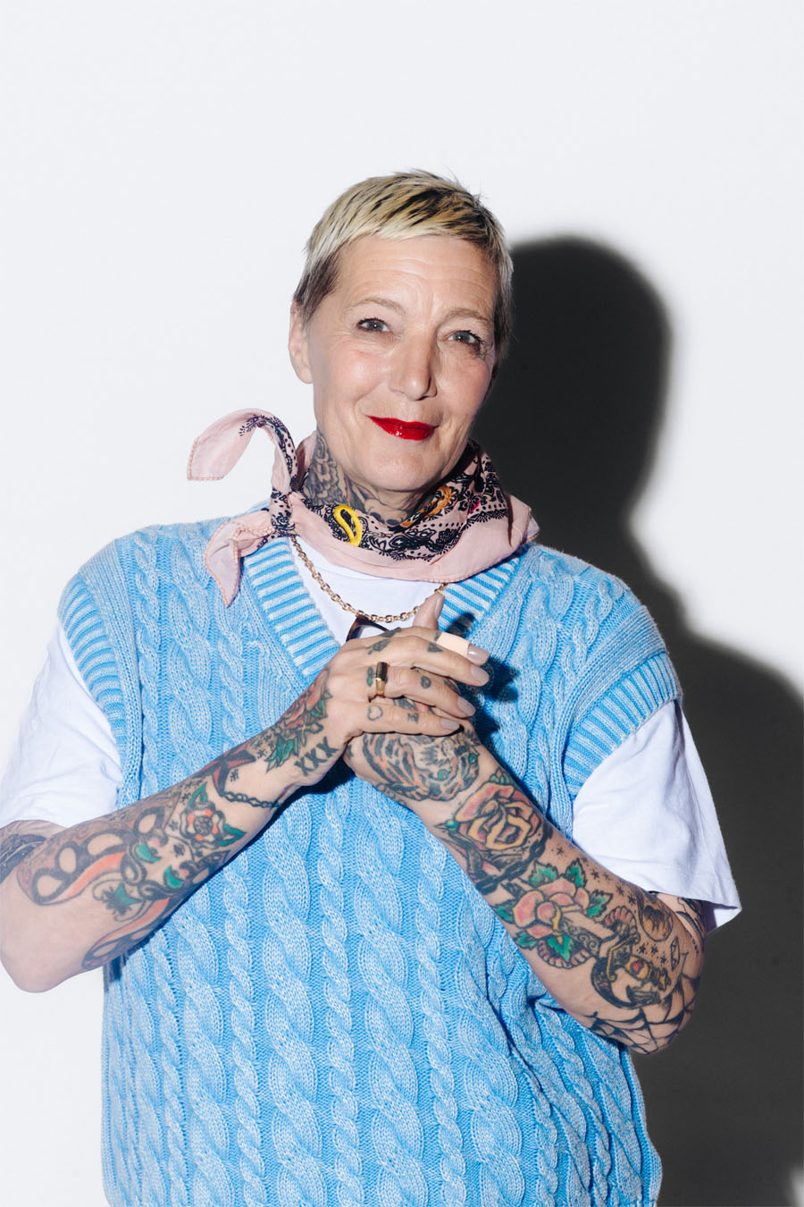 Une femme en pull bleu portant un rouge à lèvres rouge et qui a des tatouages sur les bras